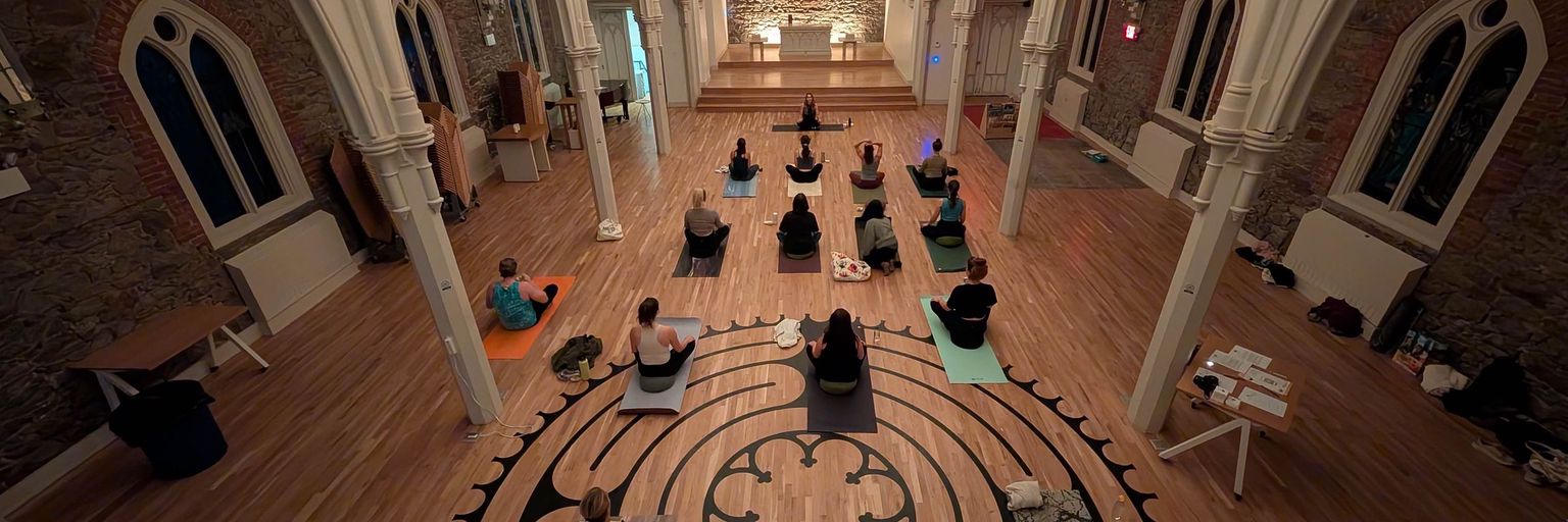 An overhead shot of a yoga class taking place within Ascension Church. Several people are sitting on yoga mats, facing the front of the church.